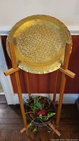 Brass wall plate with intricate lattice floral pattern and wooden plant stand visible below, with planter containing artificial greenery.