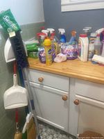Photo of various cleaning products including sprays, paper towel roll, Cascade detergent, Medix aerosol, and a broom with dustpan in the corner of the room.