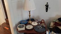 Photo showing a table with multiple clocks including crystal mantle clock, Elvis themed clock, blue and white desk clocks, brown desk clock, and a gold lamp with white lampshade.