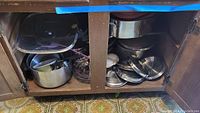 Open cabinet showing multiple pots and pans, several lids, some metal and glass, stacked in two compartments