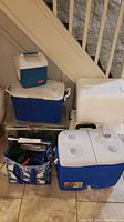 Full view of all coolers and insulated tote arranged on floor under stairs showing size variation and styles.