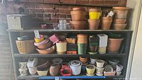 Wide view of shelves with various flower pots, planters, and garden containers, mostly terracotta and ceramic, showing weathered condition and variety of sizes.