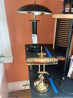 Two desk lamps on a black surface: one brass lamp with horizontal bulb case and one black lamp with dome shade and pull chain.