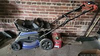 Photo of blue Kobalt KM210-06 electric lawn mower showing damaged fabric grass catcher bag, wheels, and orange power cord beside brick wall on ground.