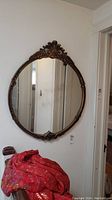 Round vintage ornate wall mirror photographed hanging on a white wall with a red cloth visible below it.