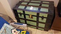Front view of vintage steamer trunk showing green and black panels with wooden slats and metal latch with padlock