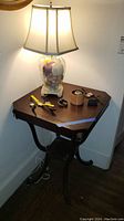 Wood accent table with curved legs supporting a glass lamp filled with seashells on top, lamp is turned on.