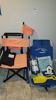 Blue Tommy Bahama chair with beach toys and signs arranged on and beside it, with orange folding director chair adjacent