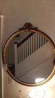 Full view of round golden enameled wall mirror hung on wall above a lamp, showing reflective surface and decorative top bow detail.