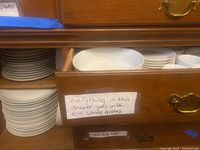 White ceramic dinner plates and salad plates stacked inside a wooden cabinet drawer with a label indicating all items in the drawer are white dishes.