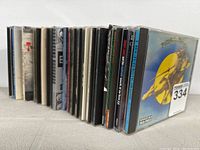 Side view of a collection of 22 jewel case CDs lined up showing the spines with artist and album names on some cases.