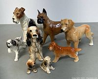 Group of seven dog figurines showing various breeds and sizes, ceramic and porcelain, with some hand paint details and a chain collar on one.