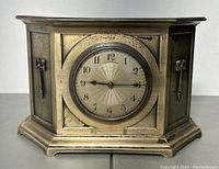 Front view of metal mantel clock showing the round face with Arabic numerals and ornate case design.