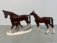 Two brown ceramic horse figurines, one adult horse with foal on base and one single standing horse, shown from side angle on white background.