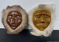 Front view of pair of Inuit style plaques showing ceramic faces with fur trim on a flat surface.