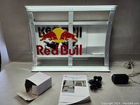 Red Bull LED lighted acrylic sign showing logo and metal frame, power adapter, mounting screws, and manual placed on a white surface.