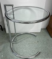 Side view of the adjustable side table showing the chrome tubular frame and clear glass top resting on a vinyl floor against a wall.