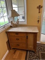 Full view of the antique pine cabinet located next to a window showing the cabinet, drawers, door, marble top, and the rooster lamp on top.