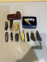 Full view of assortment of 13 pocket knives and tools on table including wood, metal and plastic handles, and a decorative knife in a tin box