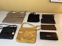 Seven various bags and wallets including beige hobo bag, black crocheted bag, gold satin bag, and assorted wallets on a white table.