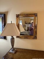 Gold framed square mirror hanging on wall above a table lamp with beige shade and wood-textured base.