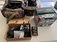 Overview photo showing the camouflage bag containing a horn speaker, a cardboard box with the Motorola walkie-talkies and accessories, a small brown Clic case, and the Coleman spotlight box.