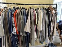 Wide view of lot showing men's clothing on hangers, mostly shirts and jackets in neutral, blue, and gray tones, various patterns including floral and stripes, mostly size XL.