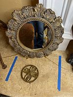 Full view of round gold ornate mirror with leaf pattern frame and brass circular floral stand on floor.