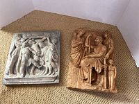 Both wall plaques shown together on carpet, grey and brown with classical scenes