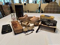 Wide view of the lot on a table showing a Charlie Chaplin cloves tin box, vintage camera in box, shoe lasts with straps, and other items.
