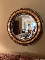 Full view of the gold round mirror hanging on a beige wall, showing its reflective surface and ornate golden frame.