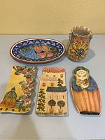 Top view of five ceramic items on table including oval fruit dish, rectangular serving dishes, sunflower vase and a figurine spoon rest.