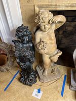 Photo showing both large angel statues side by side by fireplace on tiled floor, light statue on right holding flowers, dark statue on left holding wheat bundle.