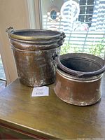 Two large copper pots placed side by side on a wooden surface by a window. Both have visible wrought iron handles and a well-worn copper exterior with tarnish and patina.