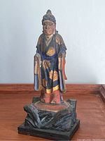 Front view of 19th century Asian wooden figure standing on a wooden carved base, dressed in a blue and red robe with gold gilt floral decoration.