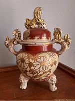 Front view of Japanese Kinkozan foo dog censer with floral patterns in cream and gold on reddish base, three foo dog legs, and foo dog figure on lid