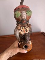 Front view of antique African figure showing large green and white bead eyes, red bead top, shell chestpiece, and wooden body with hole at base.