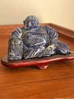 Photo showing front and left side view of the carved Hotei Buddha in blue lapis lazuli stone sitting on a wooden stand placed on wooden surface.