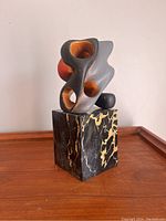 View of modernist abstract sculpture showing rounded hollow shapes and reddish-orange accents, mounted on black and gold veined marble base.