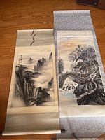 Pair of Chinese scrolls displayed on a wooden floor, one with a black and white mountainous landscape and the other with a colorful village and river scene.