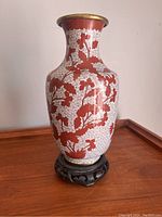 Front view of the cloisonné vase showing red floral motifs on white cloud background with brass rim and carved wooden base.