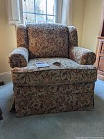 Front view of plush arm chair upholstered in floral patterned fabric, showing cushioned armrests, backrest, and skirted base.