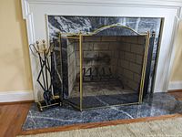 View of brass look and mesh fireplace screen next to metal fireplace tools on stand in front of a marble surround fireplace.