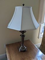 Full view of the metal table lamp on a wooden surface with white window curtain background, showing the white fabric shade and metal base.