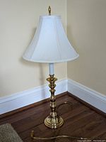 Full view of the brass look table lamp on a wooden floor against a beige wall, showing the base, pole, and white fabric shade.