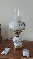 Full view of the white floral designed hurricane lamp on a wooden table, showing the glass chimney, painted flowers, and brass base.