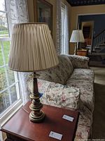 Photo of one brass look table lamp with cream pintuck fabric shade on a wooden side table near a window with floral upholstered couch in background.