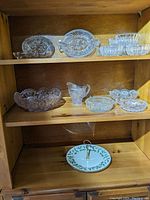 Wooden shelf holding Lenox dessert tray and various pressed glass bowls, pitcher, and relish dishes arranged across two shelves.