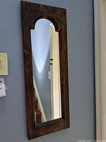 Front view of wood framed wall mirror with dark brown finish, rectangular shape and arch detail at top, showing mirror reflection and wall behind.