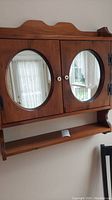 Front view of wooden cabinet with two oval mirrored doors and lower shelf, showing shape and handles.